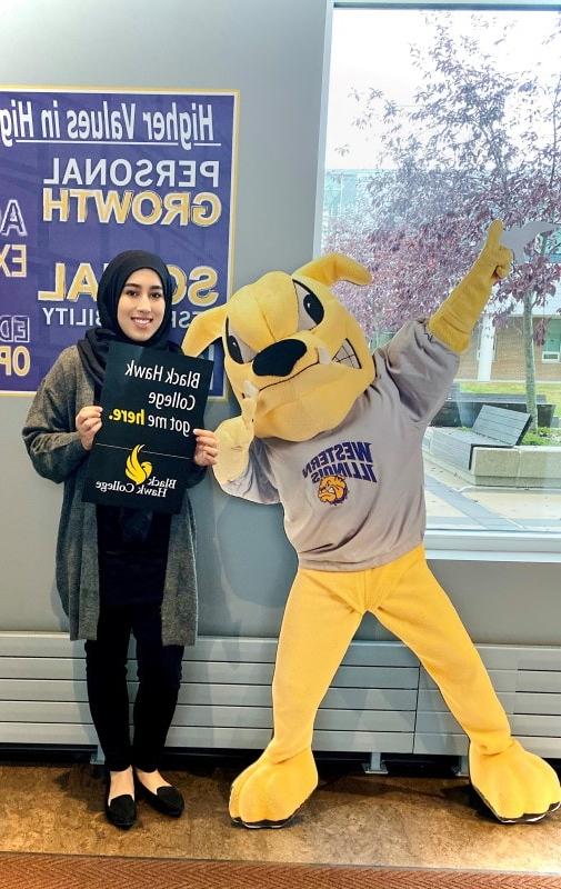 student standing next to a bulldog mascot while holding a sign that says Black Hawk College got me here